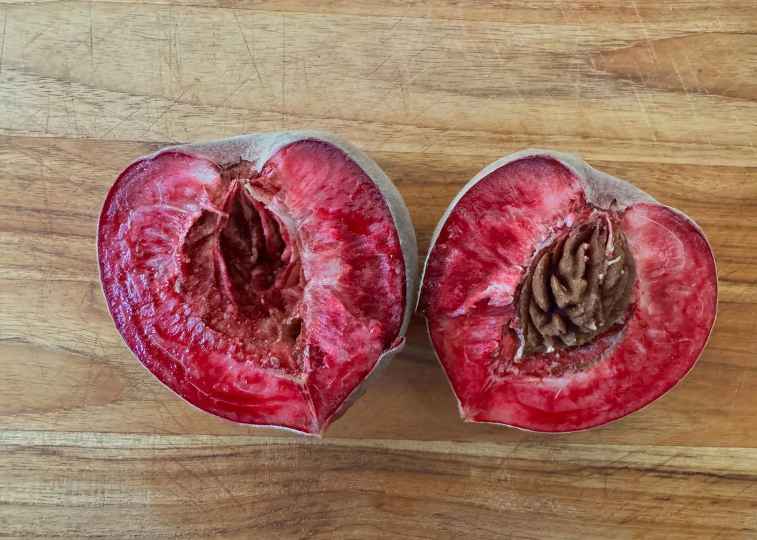 An Indian Blood peach cut in half on a wooden board, with bright red flesh and the stone still stuck in one half